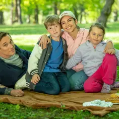Radosna rodzina na pikniku w parku. Izabela Warykiewicz z uśmiechem obejmuje dwójkę dzieci, obok siedzi mężczyzna.