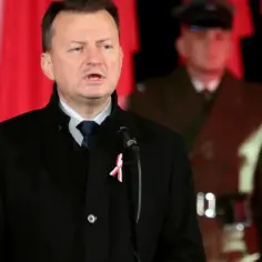 Mężczyzna w czarnym płaszczu z biało-czerwoną wstążką przemawia przy mikrofonie. W tle widać żołnierza i polskie flagi. Nie wiadomo, ile lat ma błaszczak.