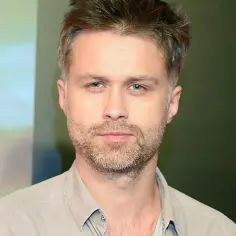 Maciej Zakościelny, aktor, z zarostem i w jasnej koszuli, na tle kolorowych paneli.