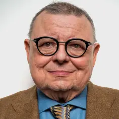 Uśmiechnięty Wojciech Mann w okularach i brązowej marynarce. Ile lat ma Wojciech Mann? Tego nie dowiemy się ze zdjęcia, ale wygląda na doświadczonego i pełnego życia.