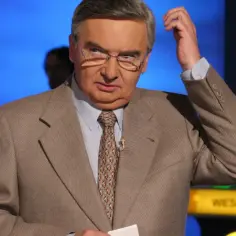 Tadeusz Szmuk, znany z teleturniejów, w garniturze i krawacie, z ręką przy głowie. Czy Tadeusz Szmuk żyje? Tak, jest aktywny.