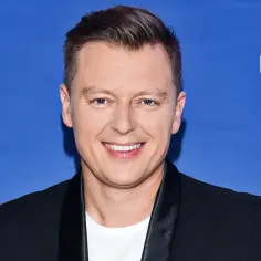 Uśmiechnięty Rafał Brzozowski na tle napisu "Bądźmy razem". Wiele osób zastanawia się, ile lat ma Rafał Brzozowski.