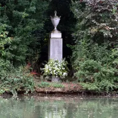 Kamienny pomnik z urną, otoczony zielenią, z bukietem białych kwiatów u podstawy. Miejsce pamięci, przypominające grób księżnej Diany.