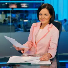 Kobieta w różowej marynarce siedzi przy biurku z dokumentami i długopisem.