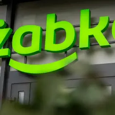 Jasnozielony neonowy napis "Żabka" z uśmiechem pod spodem na tle budynku.