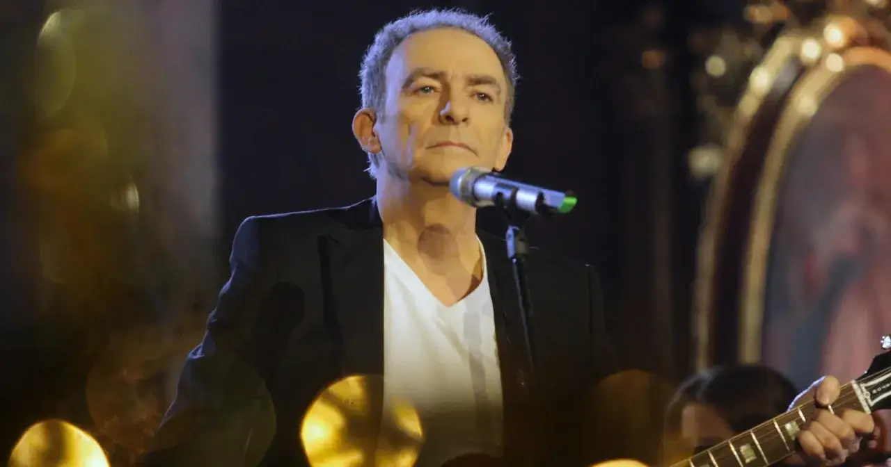 Seweryn Krajewski dzisiaj, w czarnej marynarce i białej koszulce, śpiewa przy mikrofonie, grając na gitarze.