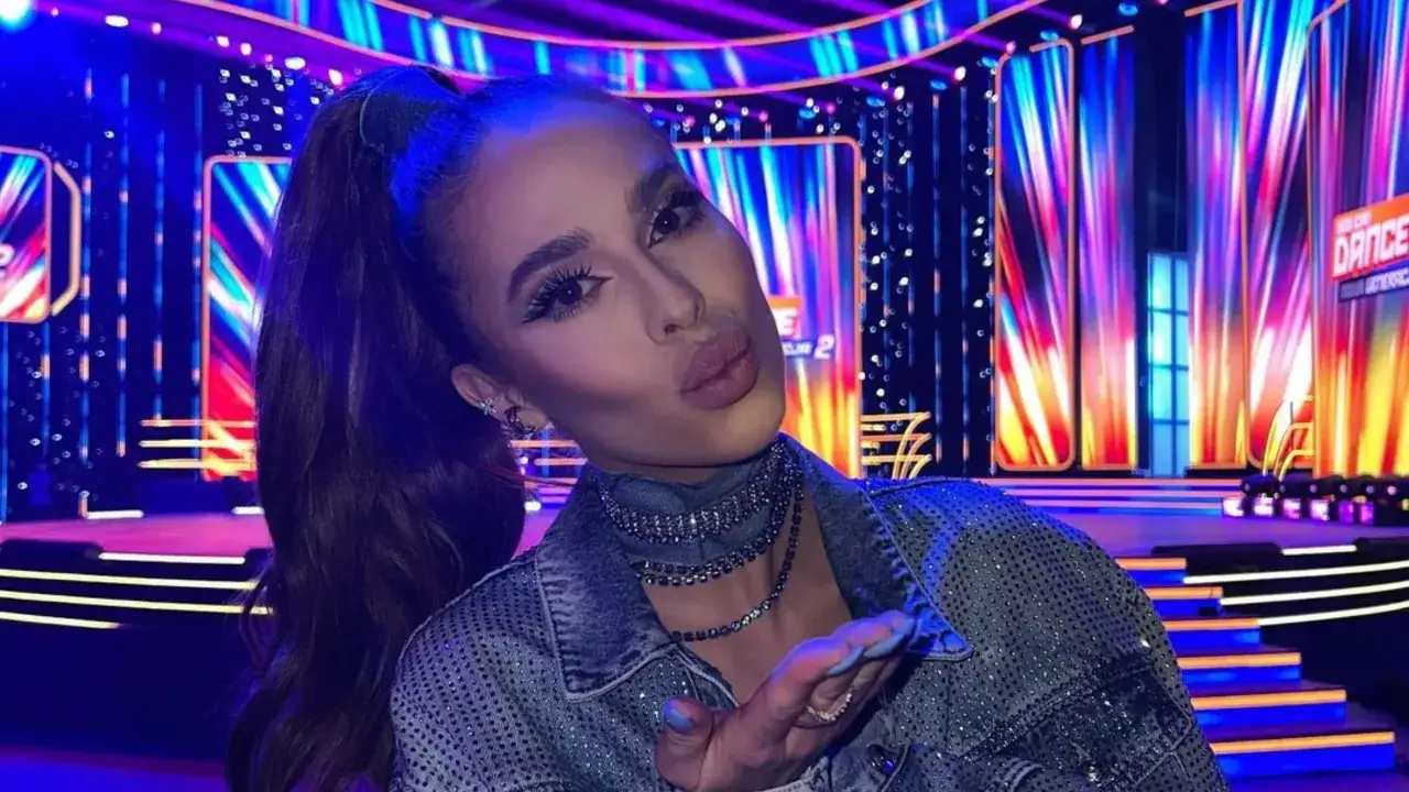 Blanka solo w błyszczącej kurtce, z wyciągniętą dłonią i ustami ułożonymi do pocałunku, na tle sceny z neonowymi światłami.