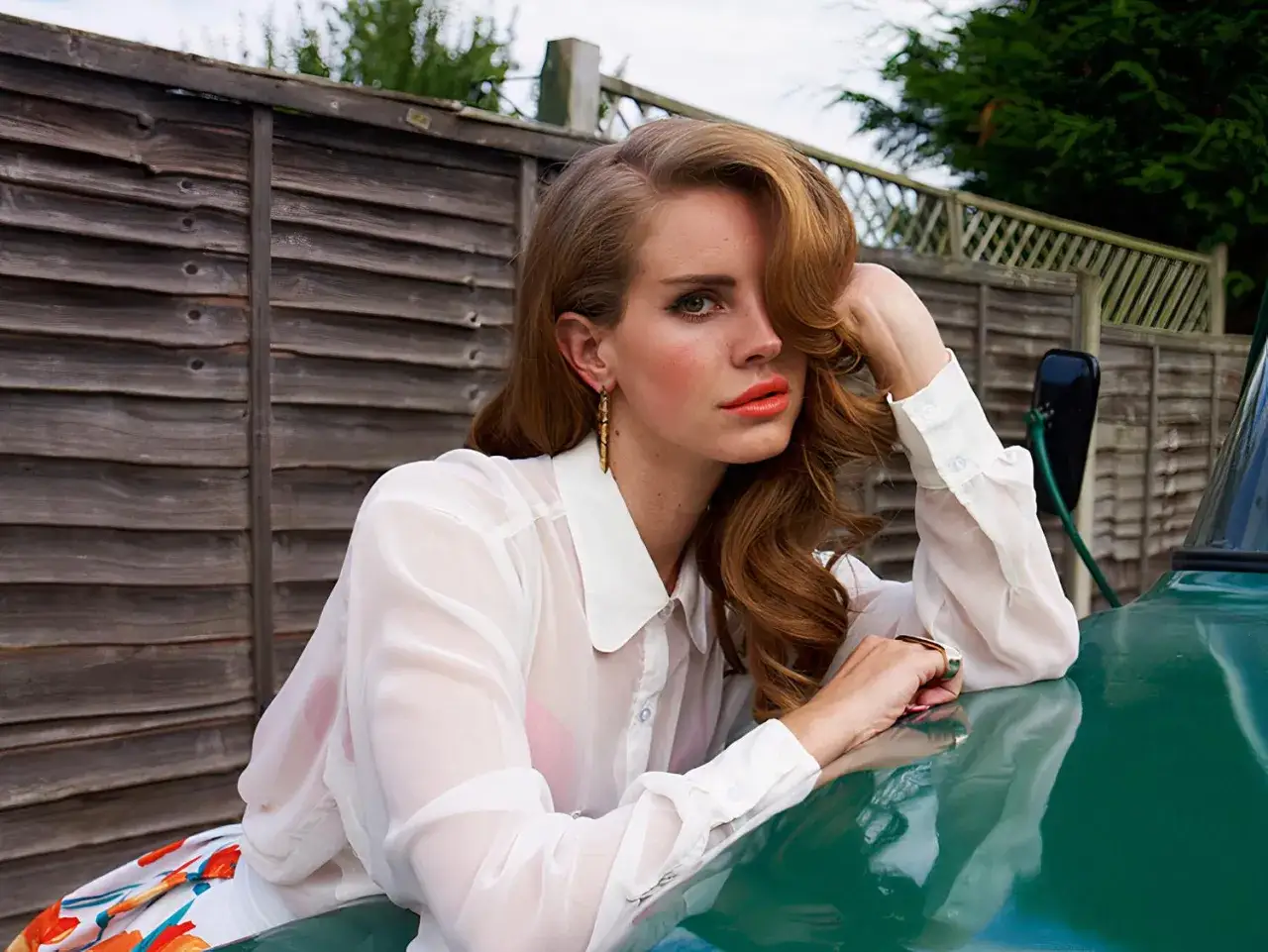 Lana Del Rey, ile lat ma Lana Del Rey? Artystka w białej koszuli opiera się o zielony samochód, za nią drewniany płot.
