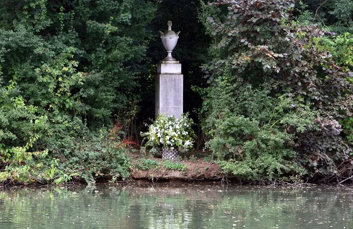 Kamienny pomnik z urną, otoczony zielenią, z bukietem białych kwiatów u podstawy. Miejsce pamięci, być może grób księżnej Diany.