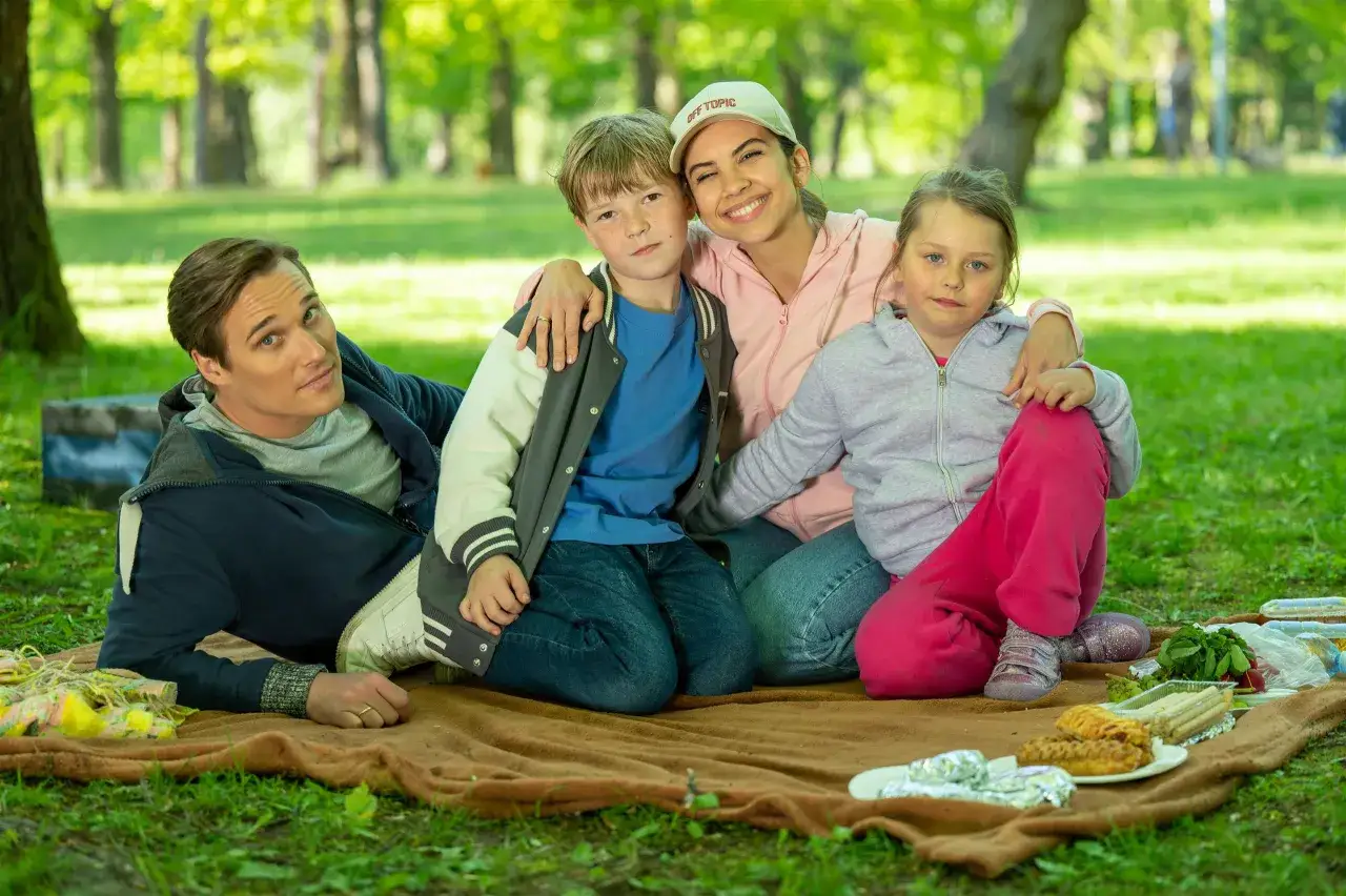 Radosna rodzina na pikniku w parku. Izabela Warykiewicz z uśmiechem obejmuje dwójkę dzieci, obok siedzi mężczyzna.