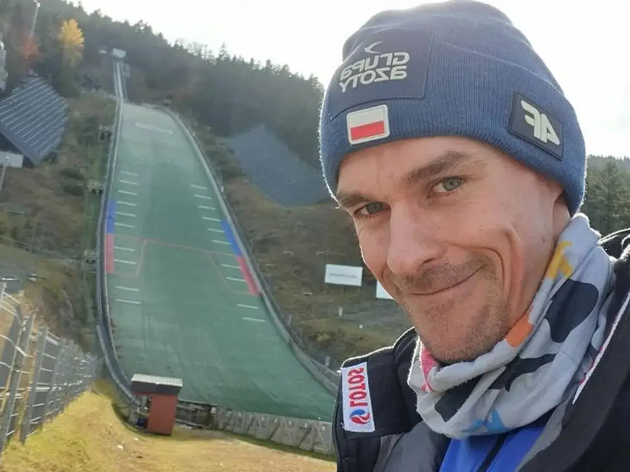 Piotr Żyła, uśmiechnięty skoczek narciarski, pozuje na tle skoczni. Ile lat ma Piotr Żyła? Tego nie wiemy, ale na pewno jest w świetnej formie!