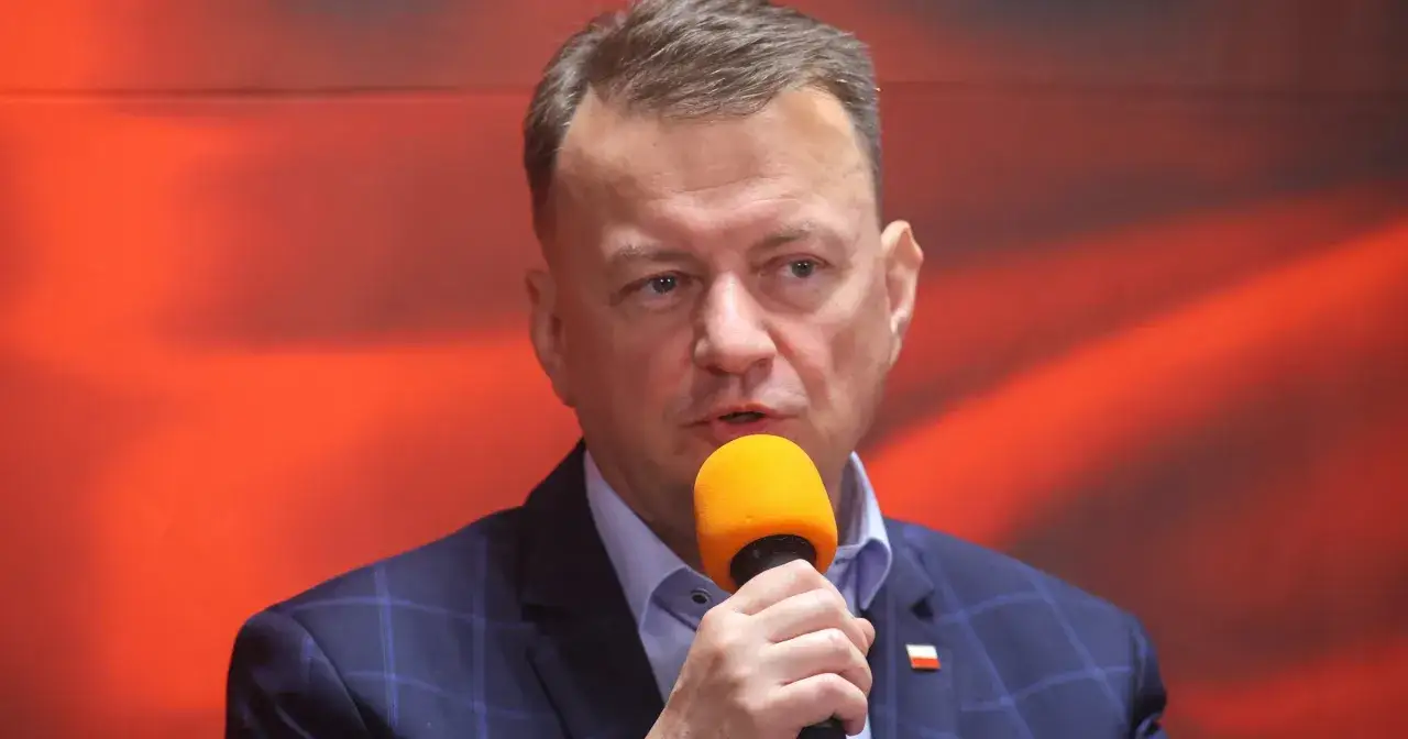 Mężczyzna w garniturze z mikrofonem, mówiący o tym, ile lat ma Błaszczak. Tło czerwone, rozmyte.
