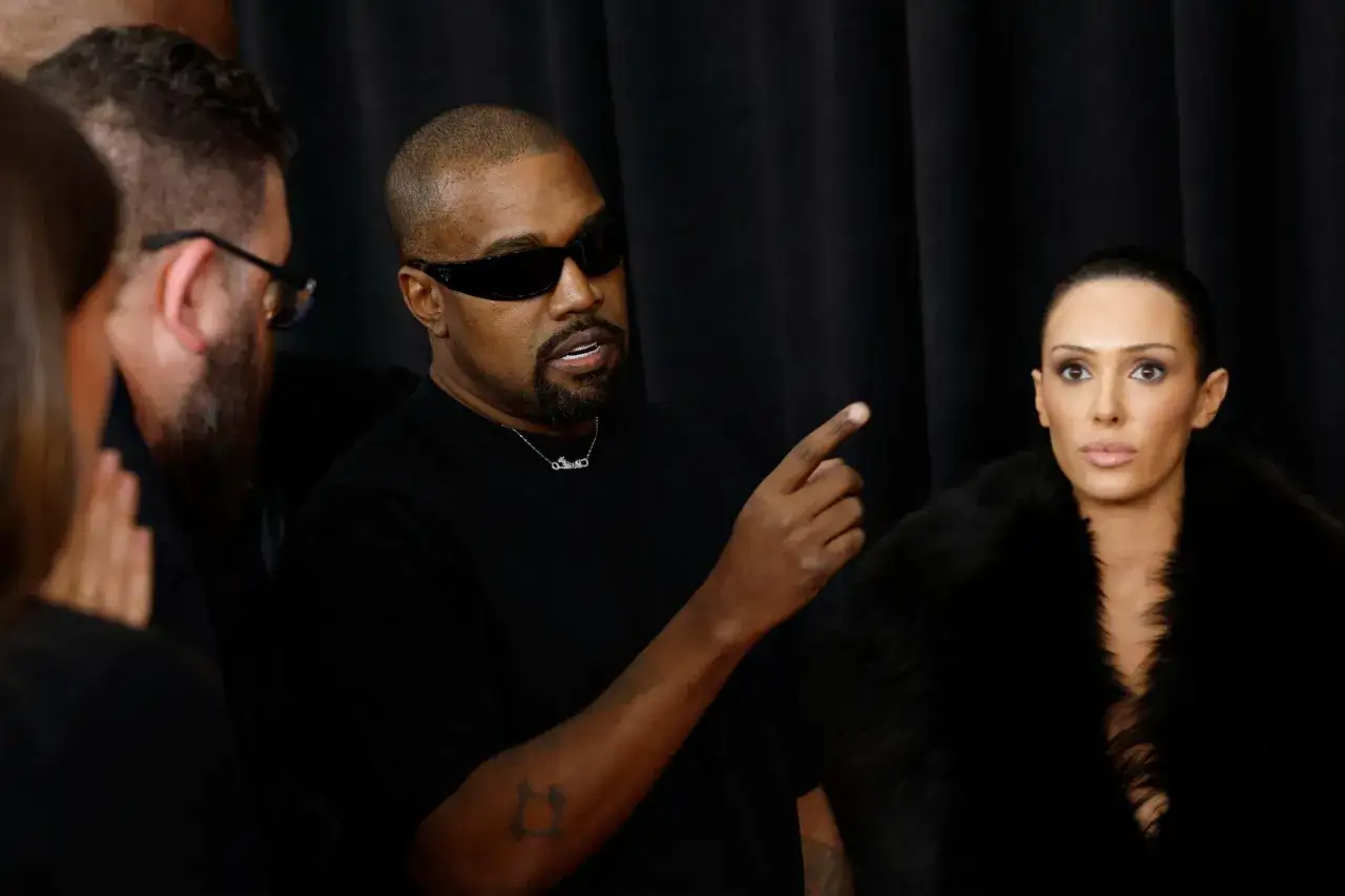 Kanye West, chory, w okularach przeciwsłonecznych i czarnym t-shircie, wskazuje palcem. Obok niego kobieta w futrzanym szalu.
